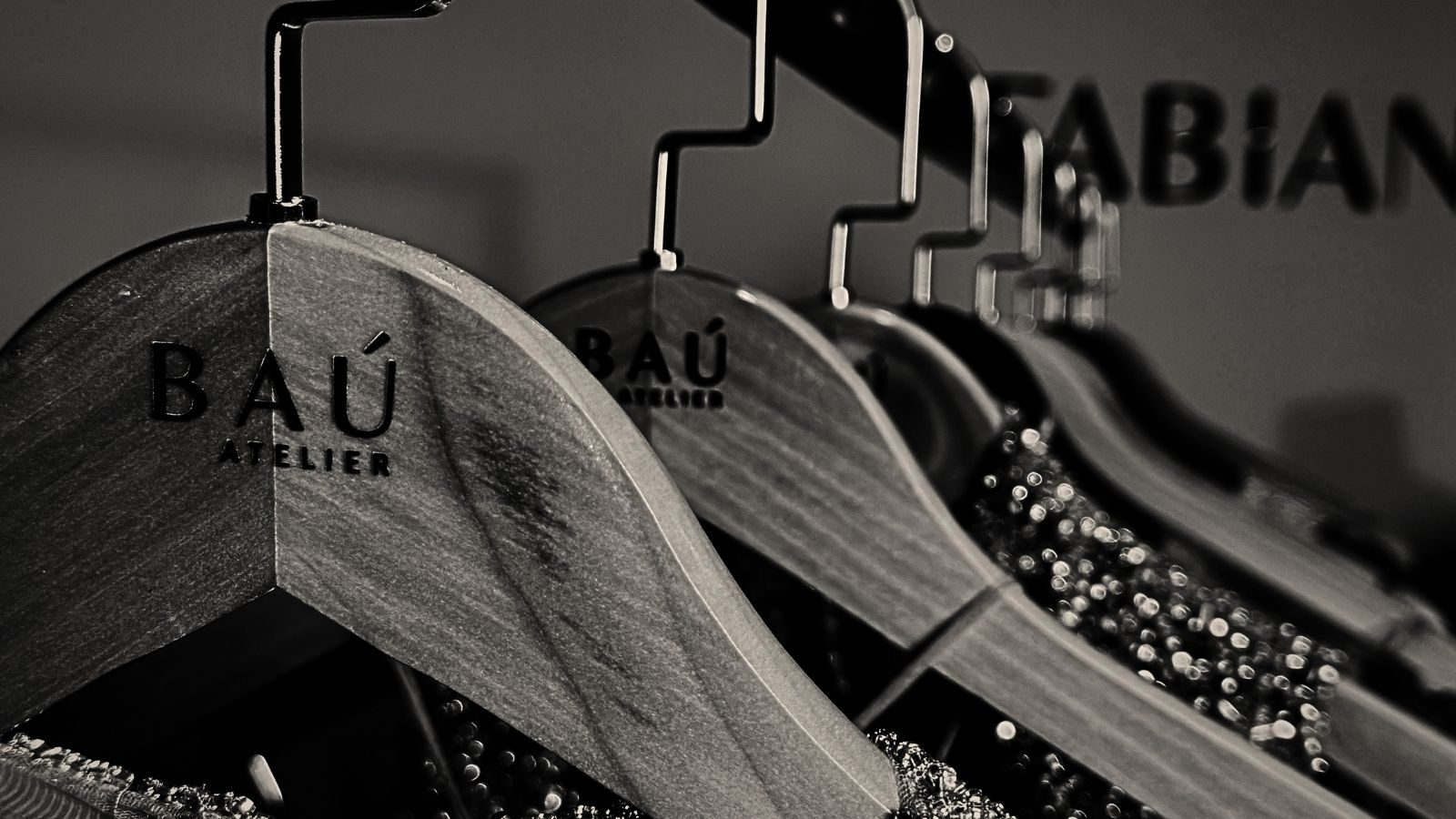 Cabides de madeira gravados BAÚ ATELIER, em fila, com vestidos bordados ao fundo