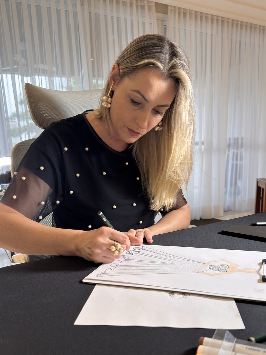 Fabiana Baú Alvarez desenhando o croqui de um vestido a lápis, na mesa do ateliê